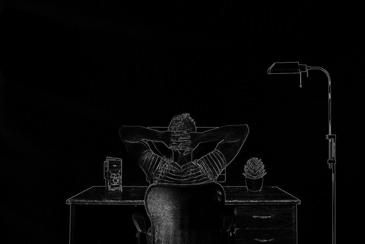 Man sitting at a desk with his hands behind his head.  His back is facing you.  Floor lamp to the right.